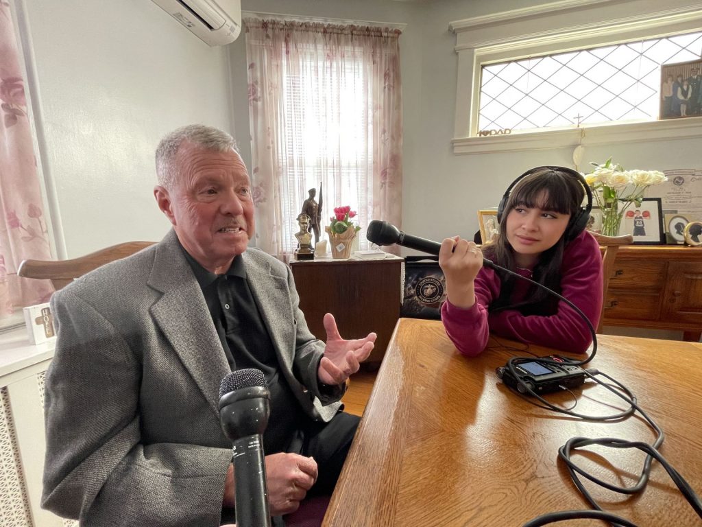 Retired NYPD inspector Charlie Wells talks and motions with his hands while being interviewed by Shoe Leather reporter Chantal Vaca, listening with headphones and holding up a microphone.