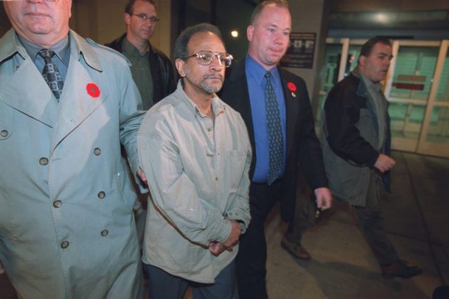 Patrick Critton walks with a stoic face and his arms crossed, escorted by police after being extradited to Canada.